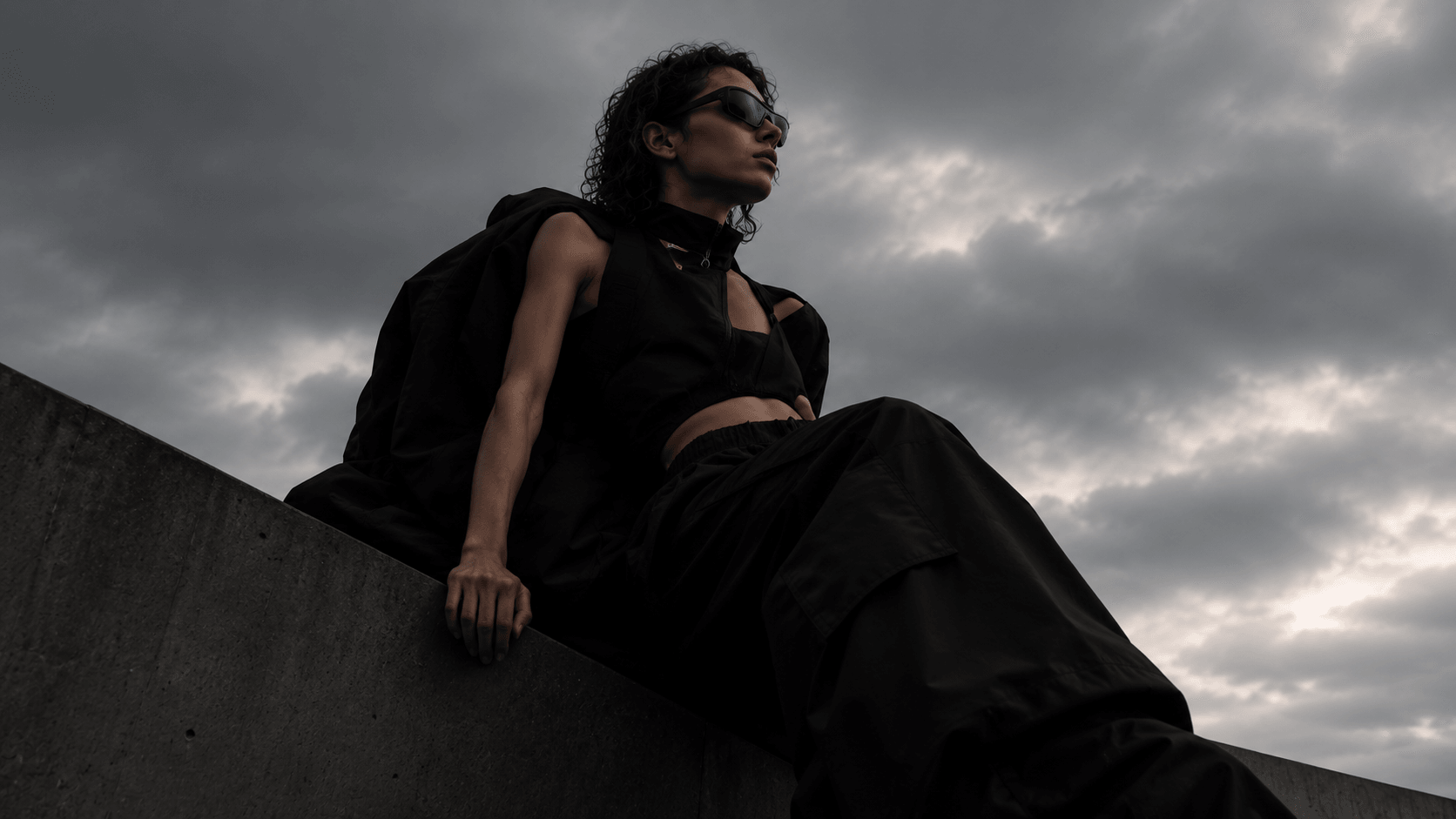 A model in black seated on a concrete ledge under an overcast sky