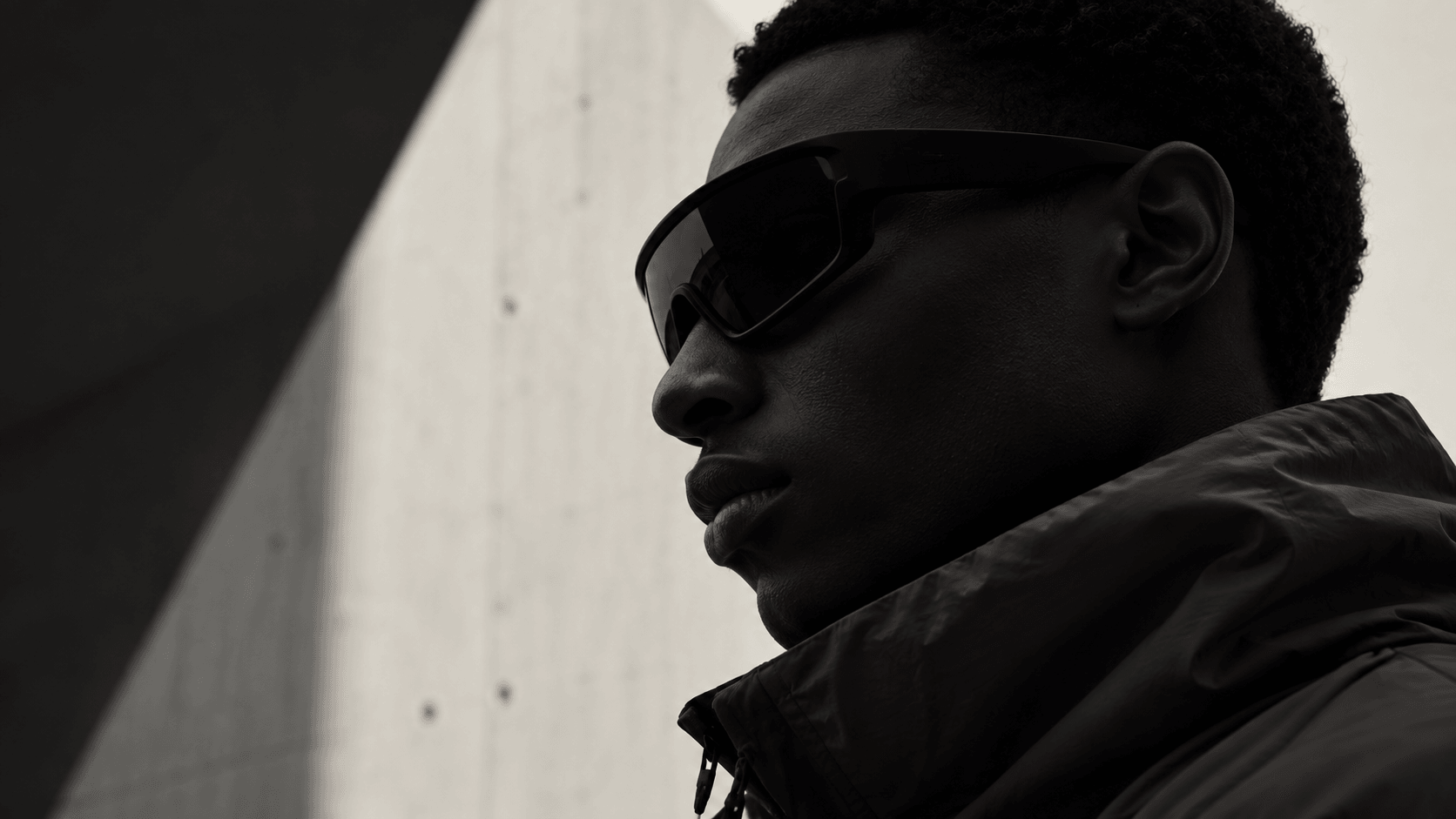 A model in wraparound sunglasses and a black shell jacket, in profile