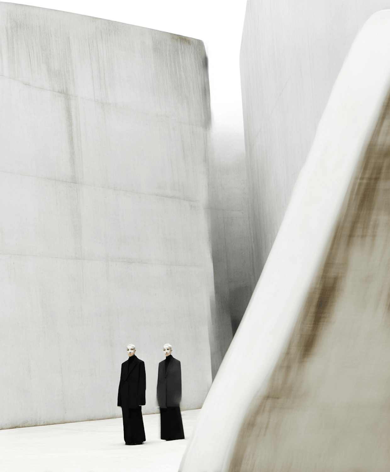 Two models in long black coats standing between tall concrete walls