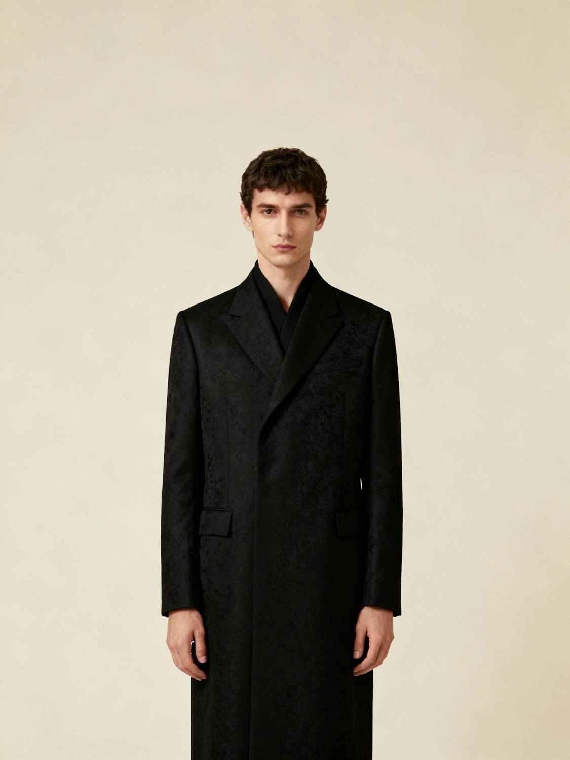 A man in a long tailored black coat against a plain wall