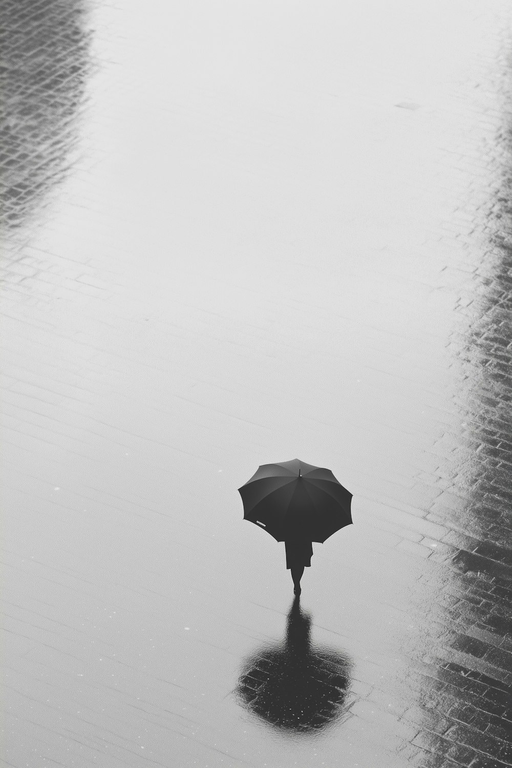 A lone figure under a black umbrella crossing wet cobblestones at night